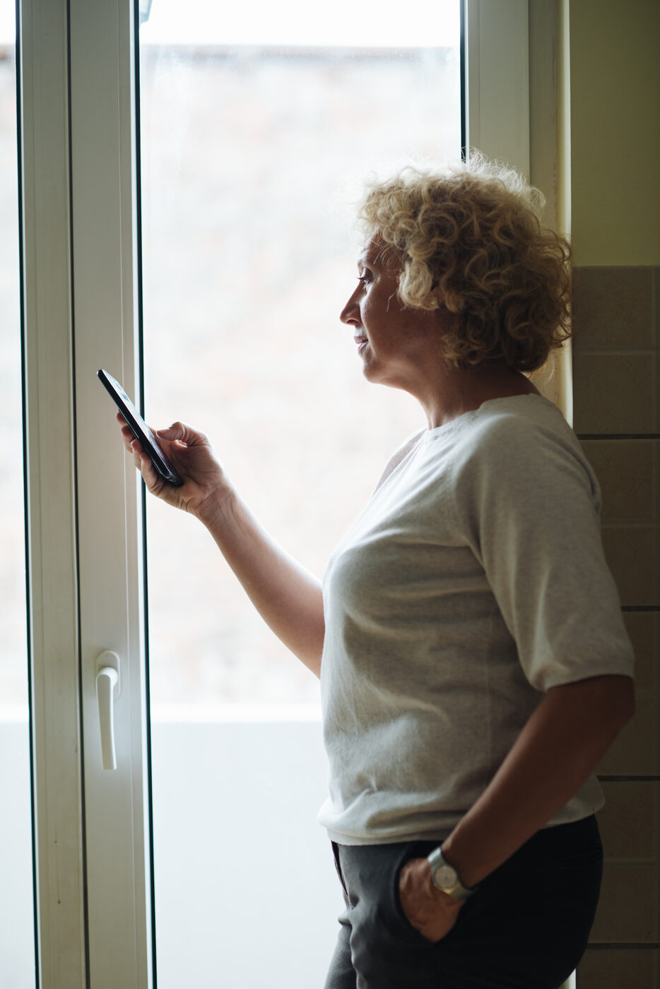 An older woman standing by a window, looking down at her phone