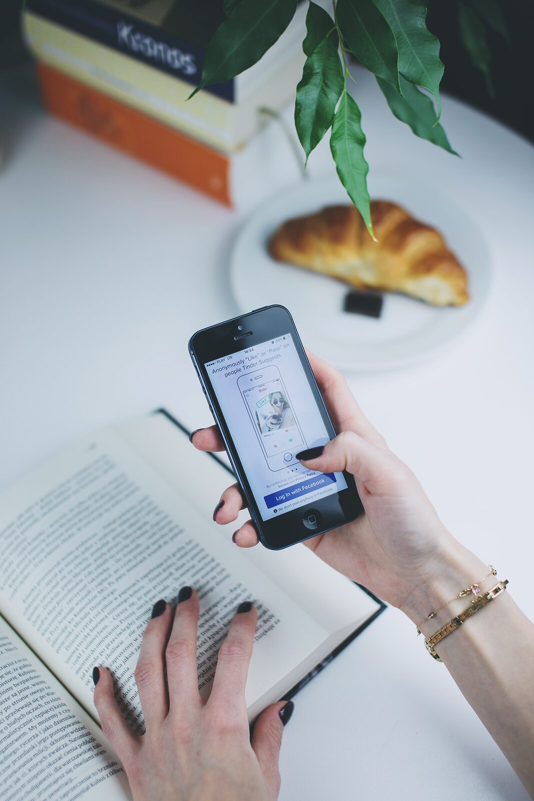 A person scrolling through Tinder on their phone while sitting at a desk with a book and a croissant — modern dating reduced to a swipe between bites