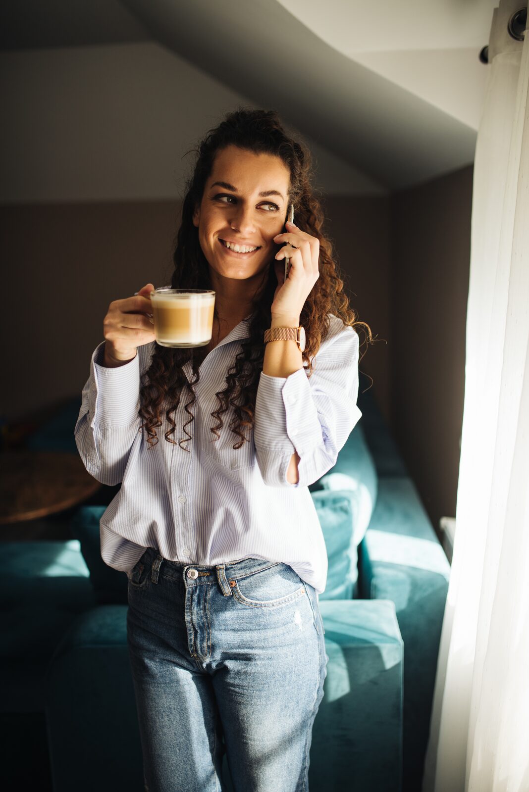 A woman holding a cup of coffee while talking on her phone in a sunlit room — the casual integration of phone use into a morning routine
