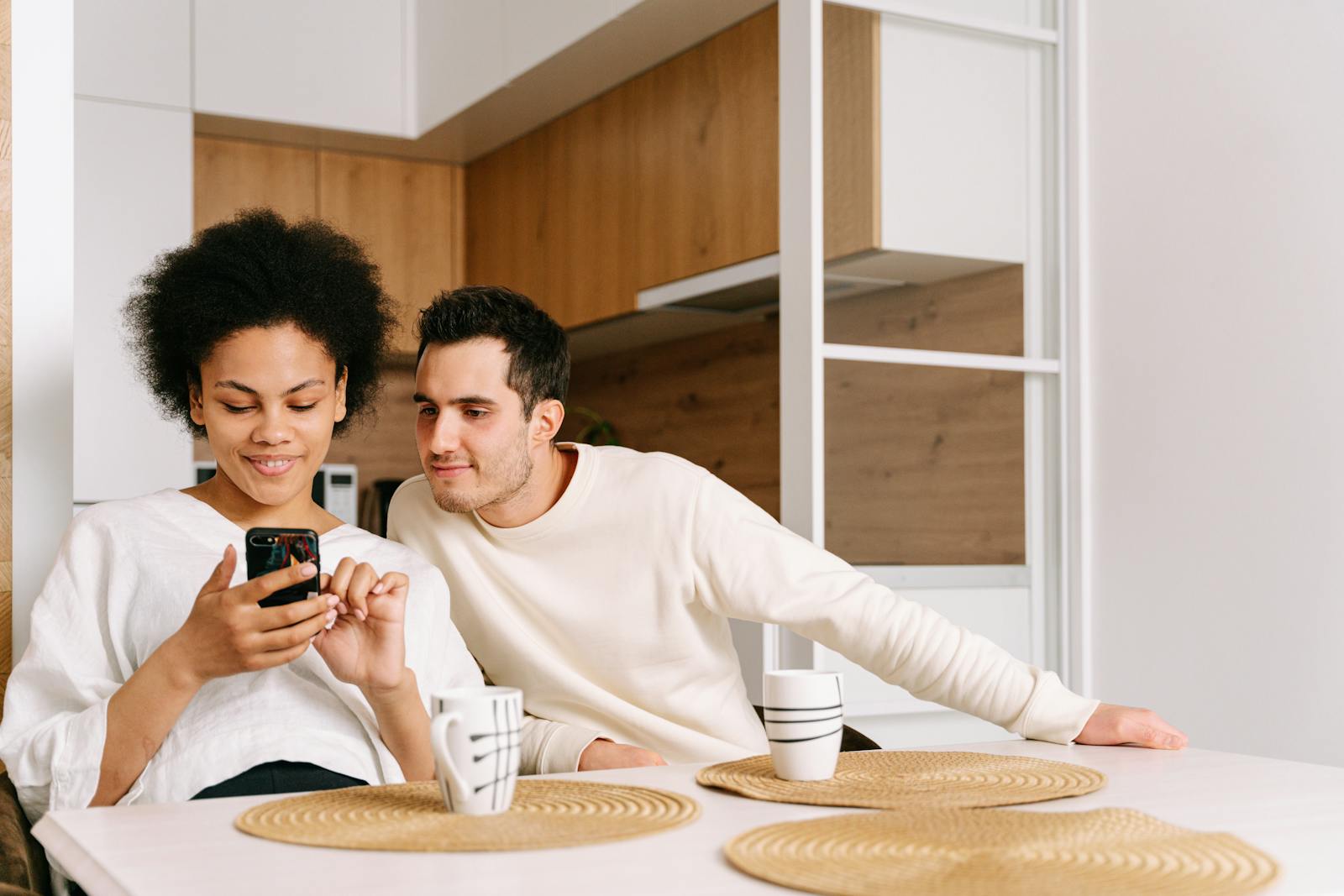 A couple sitting close together at a kitchen table, both focused on a phone screen — phones as a shared focal point in everyday relationships